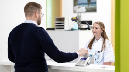 Eine Person steht am Tresen der Notaufnahme und liest eine versichertenkarte ein. Hinter dem Tresen steht eine weitere Person, die die Anmeldung in Empfang nimmt